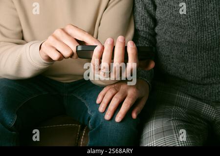 Nahaufnahme von zwei Männern in lässiger Kleidung, die nebeneinander auf einem braunen Sofa sitzen, und einer von ihnen zeigt seinem Partner etwas in seinem Smartphone Stockfoto