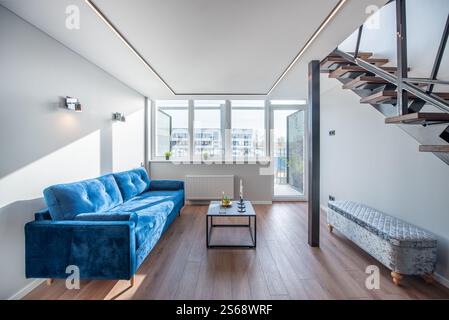 Helles und modernes Wohnzimmer mit stilvollem blauem Sofa, moderner Treppe, Holzfußboden und großen Fenstern. Home Loft Living Roo Stockfoto