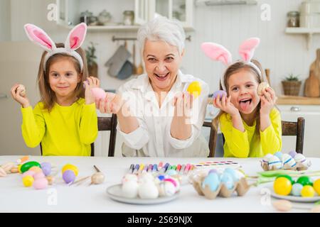 Frohe Ostern. Zwei kleine Schwestern Zwillinge, Großmutter, die Eier malt und die gemeinsame Zeit genießt. Glückliche Familie Großmutter Enkelinnen Kinder vorbereiten Stockfoto