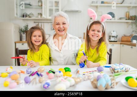 Frohe Ostern. Zwei kleine Schwestern Zwillinge, Großmutter, die Eier malt und die gemeinsame Zeit genießt. Glückliche Familie Großmutter Enkelinnen Kinder vorbereiten Stockfoto