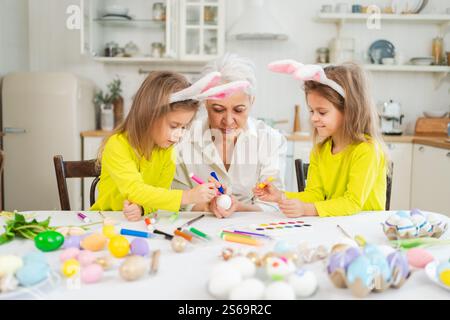 Frohe Ostern. Zwei kleine Schwestern Zwillinge, Großmutter, die Eier malt und die gemeinsame Zeit genießt. Glückliche Familie Großmutter Enkelinnen Kinder vorbereiten Stockfoto