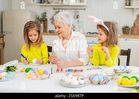 Frohe Ostern. Zwei kleine Schwestern Zwillinge, Großmutter, die Eier malt und die gemeinsame Zeit genießt. Glückliche Familie Großmutter Enkelinnen Kinder vorbereiten Stockfoto