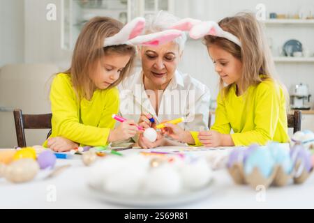 Frohe Ostern. Zwei kleine Schwestern Zwillinge, Großmutter, die Eier malt und die gemeinsame Zeit genießt. Glückliche Familie Großmutter Enkelinnen Kinder vorbereiten Stockfoto