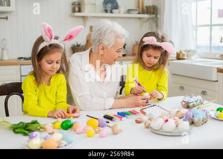 Frohe Ostern. Zwei kleine Schwestern Zwillinge, Großmutter, die Eier malt und die gemeinsame Zeit genießt. Glückliche Familie Großmutter Enkelinnen Kinder vorbereiten Stockfoto