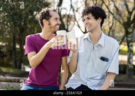 Brasilianisches Schwulenpaar tröstet mit Kaffee zum Mitnehmen, sitzt auf einer Bank im Park, lächelt und hat Spaß Stockfoto