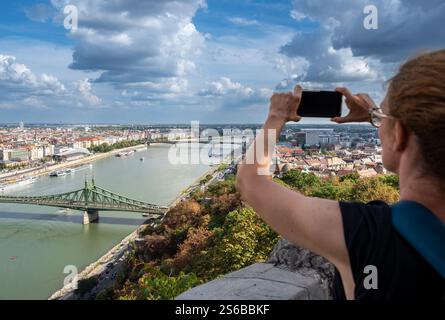 Budapest, Ungarn, 29. August 2022. Eine Frau mittleren Alters fotografiert mit ihrem Handy die herrliche Stadtlandschaft von der Spitze des Hügels. Beau Stockfoto