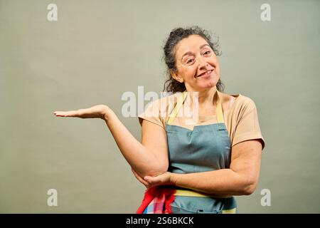 Glückliche reife Frau mit Schürze, die etwas auf der Handfläche mit Kopierraum auf neutralem Hintergrund präsentiert Stockfoto