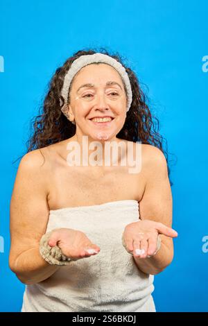Glückliche reife Frau, die ein Badetuch trägt und nach der Dusche Feuchtigkeitscreme auf ihr Gesicht aufträgt, auf blauem Hintergrund Stockfoto