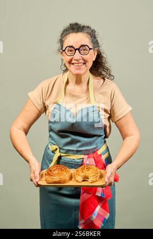 Glückliche Bäckerin in Schürze und Brille, die ein Holztablett mit zwei köstlichen, frisch gebackenen süßen Brötchen hält Stockfoto