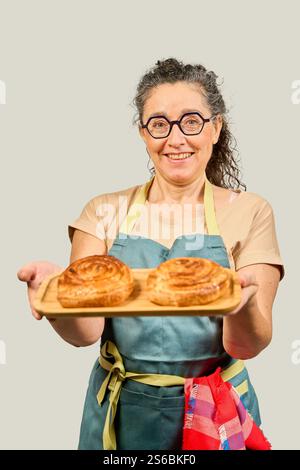 Lächelnde Bäckerin mit Schürze, die ein Holztablett mit zwei köstlichen frisch gebackenen Gebäckstücken hält Stockfoto
