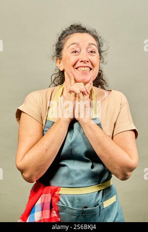 Glückliche, reife Küchenchefin trägt Schürze, hält die Hände zusammen und lächelt auf grauem Hintergrund Stockfoto