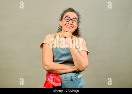 Porträt einer fröhlichen, reiferen Köchin, die Schürze und Brille trägt, Hand am Kinn hält und denkt, lächelt und in die Kamera blickt Stockfoto