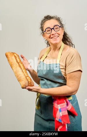 Eine Frau hält einen Laib Brot und lächelt. Sie trägt eine grüne Schürze und eine Brille Stockfoto
