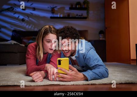 Junges Paar, das nachts mit dem Smartphone auf dem Teppich liegt und Überraschung ausdrückt. Stockfoto