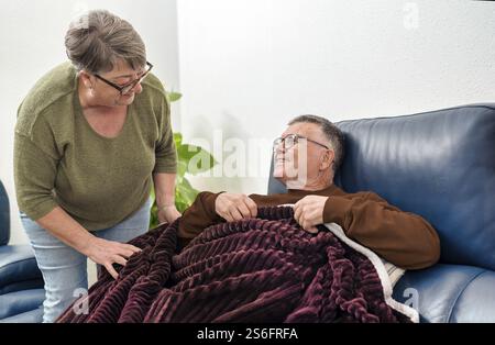 Home Healthcare Krankenschwester bietet Komfort für lächelnden älteren Mann, entspannt auf Sofa in eine warme Decke gewickelt, genießen friedliche Momente drinnen Stockfoto