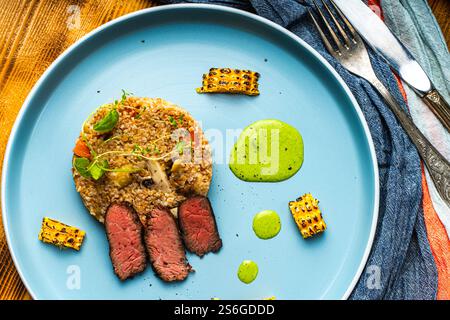 In Scheiben geschnittenes Filet Mignon Steak mit Grütze, Pilzen und Mais serviert auf einem blauen Teller mit Serviette und Besteck auf hölzernem Hintergrund. Hochwertige Fotos Stockfoto