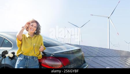 Frau mit gebogenen Haaren, die ein gelbes Hemd trägt, steht neben dem Aufladen des Elektroautos. Windräder im Hintergrund. Stockfoto