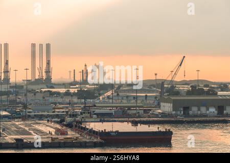 Abu Dhabi, VAE - 3. Januar 2025: Ein friedlicher Sonnenuntergang über Abu Dhabis geschäftigem Industriehafen, mit Kränen, angedockten Schiffen und Ölplattformen, die aga umrahmt Stockfoto