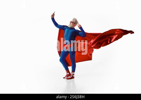 Entspannter, grauhaariger Mann im Superheldenkostüm mit rotem cape und Sonnenbrille, macht Friedenszeichen und macht Selfie vor weißem Studiohintergrund Stockfoto
