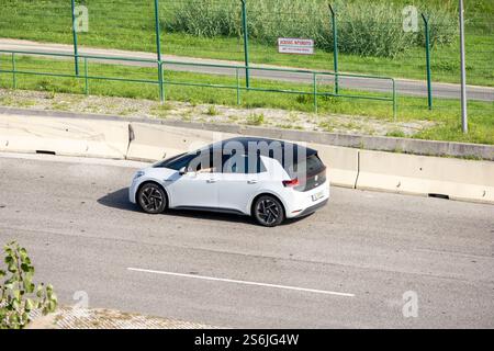Weißer Elektrowagen volkswagen ID.3, der auf der Asphaltstraße neben einem Metallzaun fährt Stockfoto