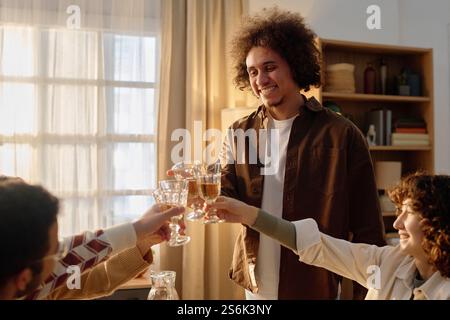 Ein kleiner Schuss des jungen Erwachsenen mit lockigen Haaren, der mit seinen Freunden eine Brille anklickt, nachdem er Toast vorgeschlagen hatte Stockfoto