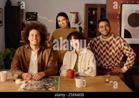 Hochwinkelporträt einer birassischen Gesellschaft von Freunden, die mit Brettspiel und Aromakerzen am Tisch sitzen und die Kamera betrachten, während sie für Gruppenfotos posieren Stockfoto