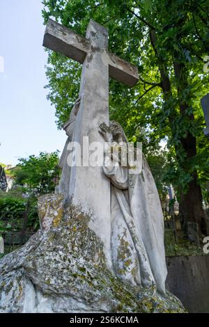 Nahaufnahme eines verwitterten Steinkreuzes mit einer moosbedeckten Trauerstatue, inmitten eines üppigen grünen Hintergrunds auf einem friedlichen Friedhof. Stockfoto