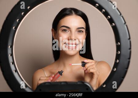 Wunderschöne junge Frau, die flüssigen Lippenstift auf beigem Hintergrund aufträgt, Blick durch Ringlampe Stockfoto