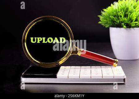 Eine Lupe hebt das Wort „UPLOAD“ auf einem Smartphone hervor, das auf einem Schreibtisch platziert ist. Der Arbeitsbereich verfügt über ein modernes Design mit einer kleinen Topfpflanze, Crea Stockfoto