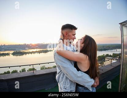 Ein junges Paar genießt einen ruhigen Abend auf einem Dach, umgeben von den warmen Farbtönen der untergehenden Sonne. Ihre Umarmung spricht von gemeinsamen Momenten und Ruhe Stockfoto