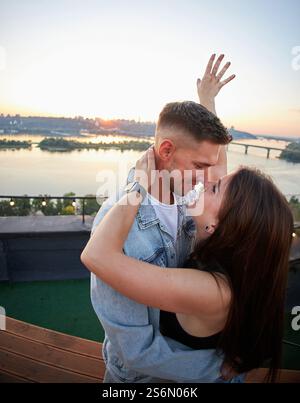 Ein junges Paar genießt einen ruhigen Abend auf einem Dach, umgeben von den warmen Farbtönen der untergehenden Sonne. Ihre Umarmung spricht von gemeinsamen Momenten und Ruhe Stockfoto