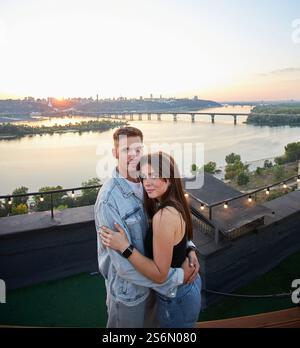 Ein junges Paar genießt einen ruhigen Abend auf einem Dach, umgeben von den warmen Farbtönen der untergehenden Sonne. Ihre Umarmung spricht von gemeinsamen Momenten und Ruhe Stockfoto