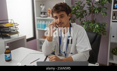 Hübscher junger hispanischer männlicher Arzt, der drinnen in einer Klinik arbeitet und mit Stethoskop um den Hals und Akten auf dem Schreibtisch telefoniert Stockfoto
