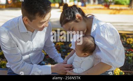 Familie genießt gemeinsame Zeit im Stadtpark, wo Mutter, Vater und Baby einen zarten Moment im Freien mit Blumen um sie herum verbringen. Stockfoto