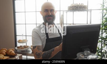 Hübscher Glatzkopf mit Bart, der in einer Bäckerei lächelt, während er eine gestreifte Schürze trägt und drinnen mit Gebäck und Kuchen um ihn herum arbeitet Stockfoto