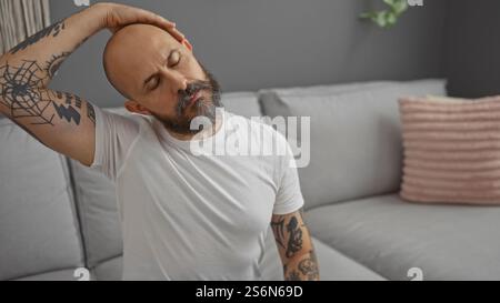 Hübscher hispanischer Mann mit Bart und Tätowierungen, die den Hals zu Hause im Wohnzimmer dehnen Stockfoto