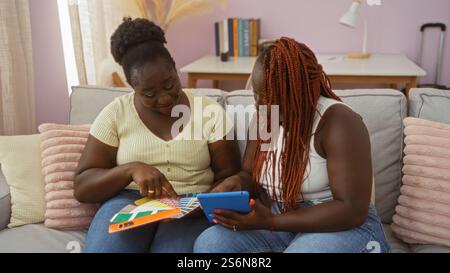 Zwei schwarze afroamerikanische Freundinnen sitzen zusammen in einem Wohnzimmer und genießen einen gemütlichen Moment, indem sie Farben aus einer Palette auswählen, während sie ein Tablet verwenden. Stockfoto