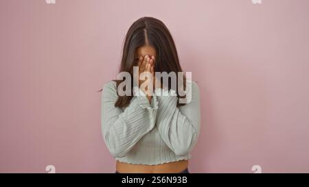 Frau mit brünetten Haaren in einem hellen Pullover bedeckt das Gesicht mit Händen, die vor einem rosa Wandhintergrund stehen und Schönheit und Emotionen in einem isolierten s ausstrahlen Stockfoto