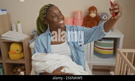 Frau lächelt drinnen und hält Ihr Baby, während sie im Schlafzimmer Selfie macht, mit fröhlichem Ausdruck und gemütlicher Atmosphäre mit einer Wiege in der Nähe. Stockfoto