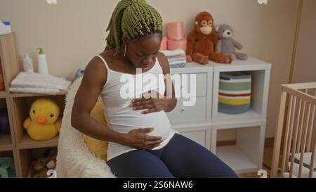 Frau mit Zöpfen sitzt in einem Schlafzimmer und berührt ihren Bauch, umgeben von Babyartikeln in einer gemütlichen Wohnumgebung. Stockfoto