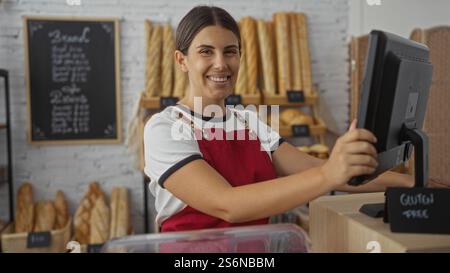 Eine junge Frau mit brünetten Haaren arbeitet in einer Bäckerei mit roter Schürze, umgeben von verschiedenen Brotsorten in einem Innengeschäft. Stockfoto