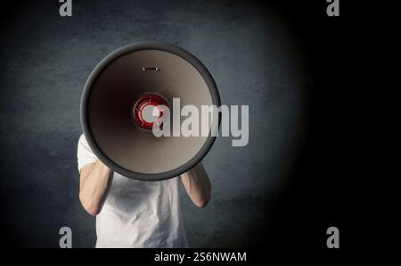 Ein junger Mann hält ein großes Megafon und spricht laut auf einem dunklen Studiohintergrund mit Platz für Ihren Text oder Ihr Bild Stockfoto