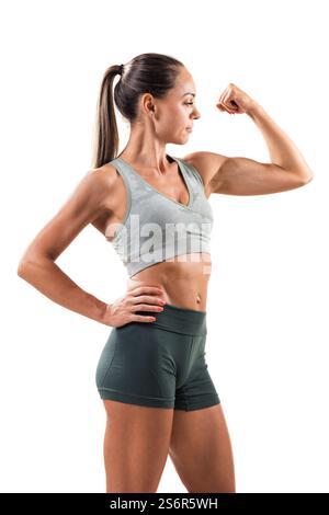 Sportliche junge Frau in Trainingskleidung, die ihren Bizeps beugt, muskuläre Definition und Selbstvertrauen zeigt. Der isolierte Studiohintergrund betont Stockfoto