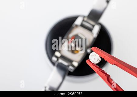 Pinzette mit Uhrenbatterie, Uhrengehäuse im Hintergrund, Unschärfe, Detail, weißer Hintergrund, Symbolfoto, Batteriewechsel, Uhrenreparatur, Stockfoto