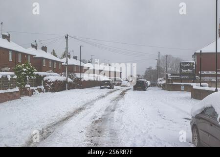 Schneebedeckte Wohnstraße mit geparkten Autos, Backsteinhäusern und einem Topps Tiles Schild. Reifenspuren sind auf der schneebedeckten Straße in Harrogate sichtbar. Stockfoto