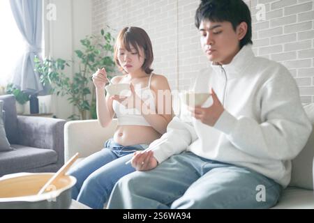 Eine schwangere japanische Frau im Alter von 20 Jahren und ihr Mann, gekleidet, um ihre Mägen zu zeigen, sitzen auf einem Sofa in einer Wohnung. Stockfoto