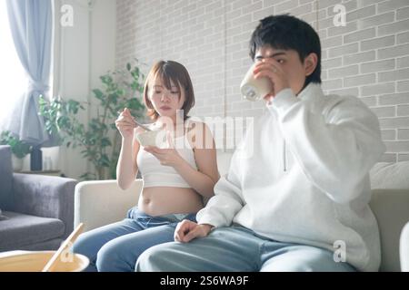 Eine schwangere japanische Frau im Alter von 20 Jahren und ihr Mann, gekleidet, um ihre Mägen zu zeigen, sitzen auf einem Sofa in einer Wohnung. Stockfoto