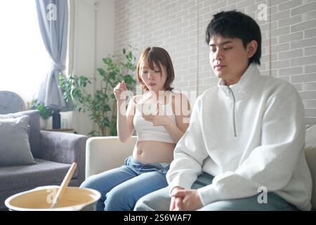 Eine schwangere japanische Frau im Alter von 20 Jahren und ihr Mann, gekleidet, um ihre Mägen zu zeigen, sitzen auf einem Sofa in einer Wohnung. Stockfoto