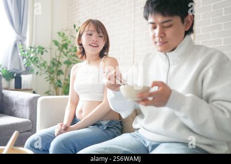 Eine schwangere japanische Frau im Alter von 20 Jahren und ihr Mann, gekleidet, um ihre Mägen zu zeigen, sitzen auf einem Sofa in einer Wohnung. Stockfoto