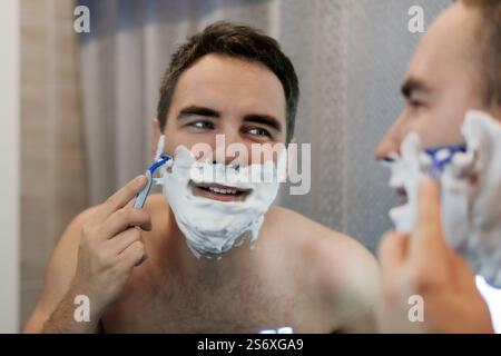 Gutaussehender, lächelnder junger Mann, der im Badezimmer mit einem Handtuch auf der Schulter steht, Rasiermesser hält und in den Spiegel blickt, mit der morgendlichen Routine beschäftigt, fürsorglich Stockfoto